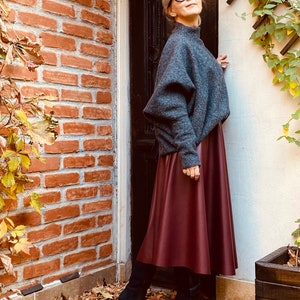 May include: A woman wearing a gray sweater, a burgundy midi skirt, and black high-heeled boots. She is also wearing sunglasses. The outfit is set against a backdrop of a brick wall and a door.