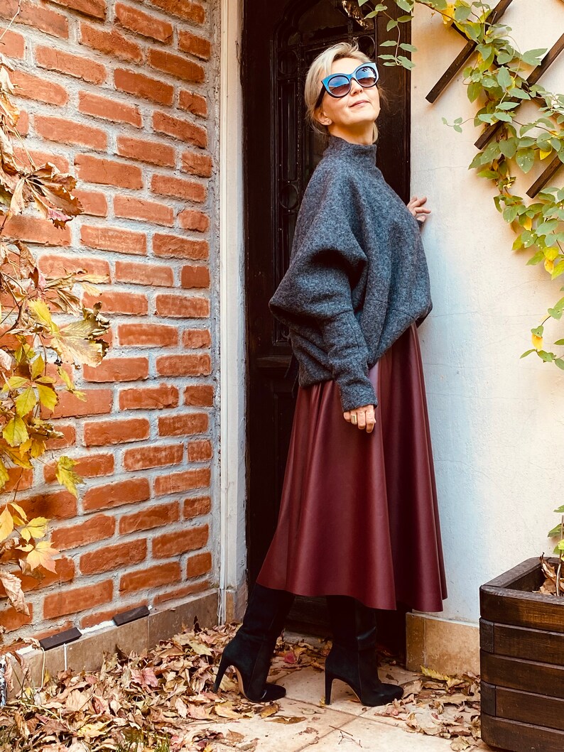 May include: A woman wearing a gray sweater, a burgundy midi skirt, and black high-heeled boots. She is also wearing sunglasses. The outfit is set against a backdrop of a brick wall and a door.