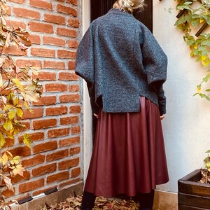 May include: A gray wool jacket with wide sleeves and a burgundy leather skirt. The outfit is paired with black high-heeled boots. The background features a brick wall and autumn foliage.