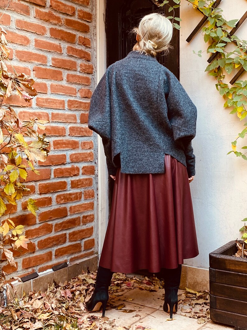 May include: A gray wool jacket with wide sleeves and a burgundy leather skirt. The outfit is paired with black high-heeled boots. The background features a brick wall and autumn foliage.