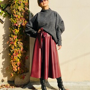 May include: A woman wearing a gray sweater and a burgundy faux leather midi skirt. The skirt is tied at the waist with a matching belt. She is also wearing black high-heeled boots and sunglasses. The background is a beige wall with climbing plants.