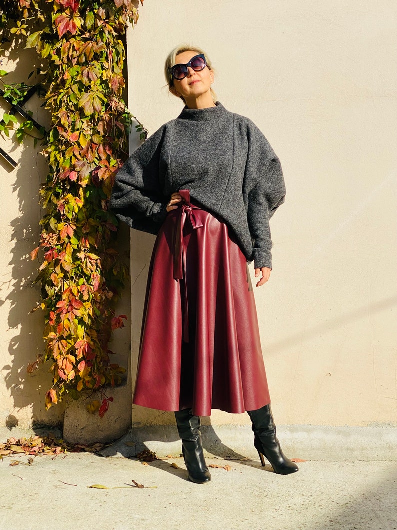 May include: A woman wearing a gray sweater and a burgundy faux leather midi skirt. The skirt is tied at the waist with a matching belt. She is also wearing black high-heeled boots and sunglasses. The background is a beige wall with climbing plants.