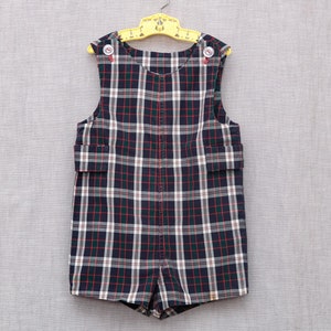 May include: A black and white plaid overall romper with red accents. The romper has a sleeveless design and is hanging on a yellow hanger.