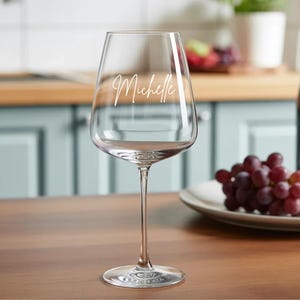 May include: A clear wine glass with the name "Michelle" in cursive script. The glass is on a wooden surface, with a plate of grapes and a bottle of wine in the background. The glass has a long stem.