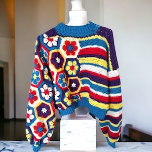 Könnte beinhalten: Ein bunter Häkelpullover mit einem geometrischen Muster in Rot-, Gelb-, Blau-, Weiß- und Lilatönen. Der Pullover hat ein gestreiftes Muster an den Ärmeln und ein gehäkeltes Blumenmuster auf der Vorderseite.