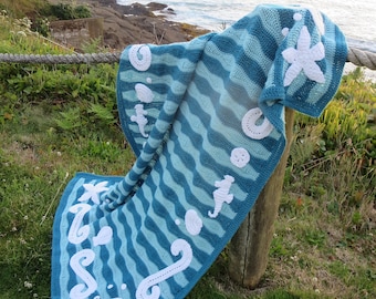 May include: A blue and teal crocheted blanket with white crocheted sea life designs, including a starfish, a seahorse, and a seashell. The blanket is draped over a wooden fence post with a view of the ocean in the background.