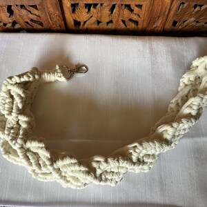 Op de afbeelding: Crèmekleurige macramé tasriem met een zilverkleurige sluiting. De riem is gemaakt van dik, geweven katoenen koord, waardoor een getextureerd, gevlochten ontwerp ontstaat. De riem ligt op een wit oppervlak.