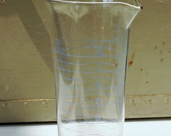 May include: A clear glass graduated cylinder with a wide base and a spout. The cylinder is marked with blue lines and numbers indicating volume in milliliters.