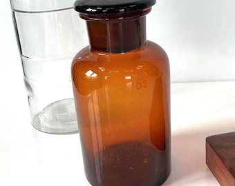 May include: An amber glass apothecary bottle with a matching dark brown glass stopper, featuring the number 02 etched on both the bottle neck and the stopper. A clear glass vessel is positioned behind the amber bottle in the background.