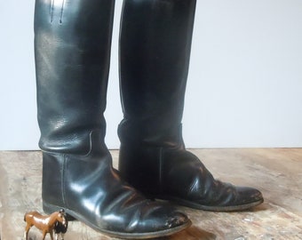 May include: A pair of black leather riding boots with a pointed toe and a zip at the back. The boots are on a wooden surface.