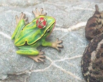 May include: A green and yellow frog figurine with a red and white hat sits on a gray rock next to a brown toad.