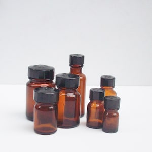 May include: A set of seven amber glass bottles with black screw-on lids. The bottles are of varying sizes and are arranged in a row on a white surface.