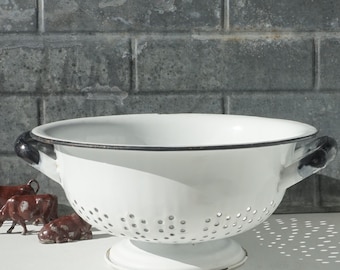 May include: White enamel colander with black rim and handles. The colander has a round base and a perforated bowl.