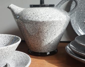 May include: A grey speckled ceramic teapot with a black lid and base. The teapot has a curved handle and spout. A matching bowl and saucer are visible, along with a stack of plates.