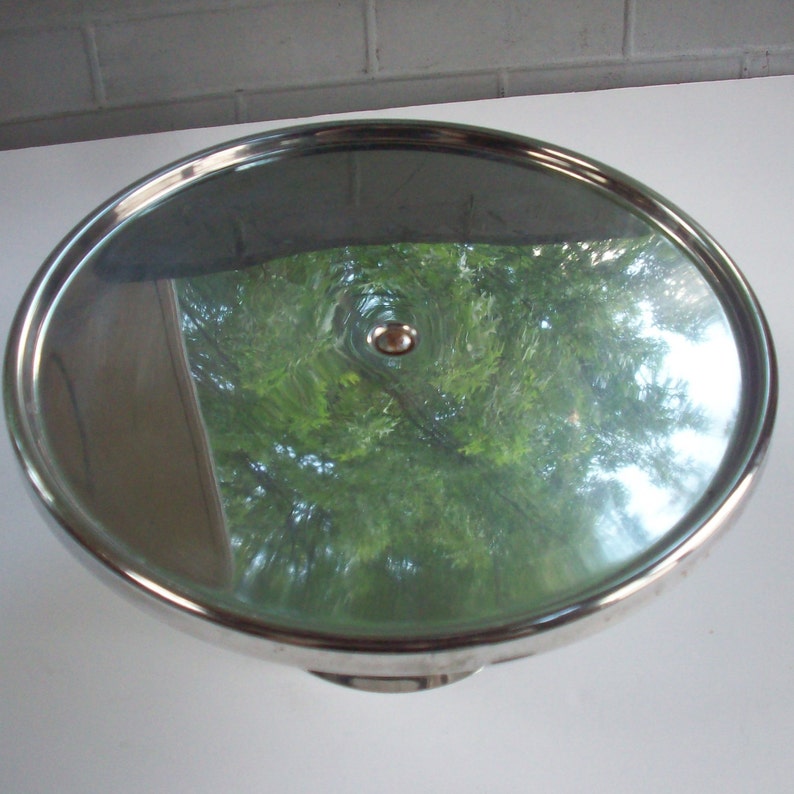 May include: A silver cake stand with a round, reflective surface and a small knob in the center. The stand has three legs.