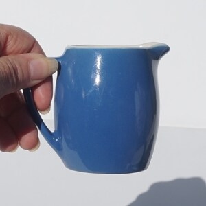 May include: A small, blue ceramic creamer with a white rim. The creamer has a rounded body and a small handle.