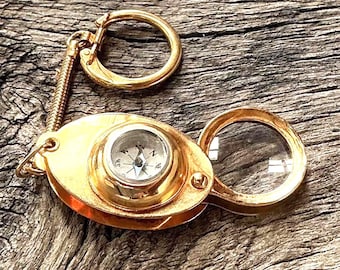 May include: A vintage-style gold-colored compass and magnifying glass keychain. The compass features a detailed dial, and the magnifying glass is open. The keychain is attached to a gold-colored spring and a large ring.