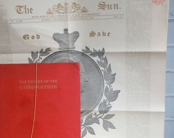 May include: A black and white newspaper with the title "The Sun." The newspaper is folded in half and has a red book titled "The History of the Coronation" on top of it. The newspaper has a royal crest and the words "God Save" printed on it.