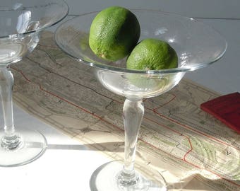 May include: Two clear glass stemmed serving dishes, each holding a lime. The dishes have a wide, shallow bowl and a slender stem. The limes are bright green. The dishes are on a map.