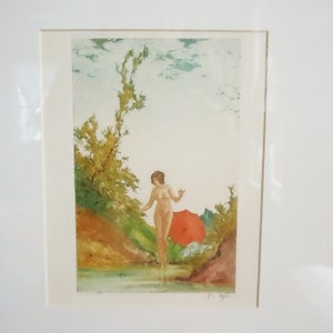 May include: A vintage print of a nude woman standing in a stream with a red umbrella in the background. The woman is surrounded by green trees and bushes. The print is framed in a white mat.