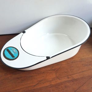 May include: White enamelware basin with a black trim and a rounded shape. The basin has a blue and black label that reads "JONES". The basin is on a wooden surface.