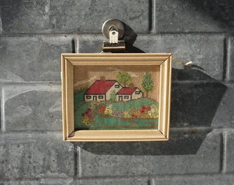 May include: A framed cross-stitch of a countryside scene with two houses with red roofs and green trees. The houses are surrounded by a field of flowers.