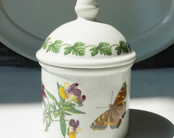 May include: White ceramic jar with a lid, featuring a butterfly and floral design. The jar has a green leafy border around the lid and a floral design with a butterfly on the body. The jar is decorated with a butterfly and flowers in shades of purple, yellow, and green.