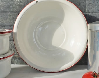 May include: A large, white enamel bowl with a red rim. The bowl is empty and has a small hole near the top edge. Other white enamel containers with red rims are visible in the background, along with a red tomato.