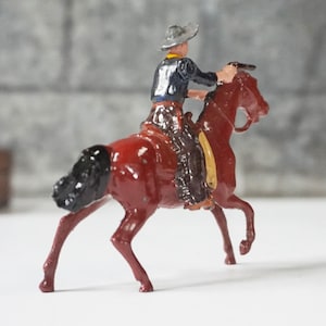 May include: A vintage toy cowboy riding a red horse with a black mane and tail. The cowboy is wearing a blue shirt, brown pants, and a brown hat with a yellow band.
