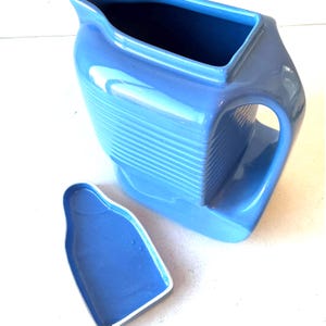 May include: A bright blue ceramic pitcher with a ribbed texture on the side and a matching blue lid placed separately. The pitcher features a wide pouring spout and a sturdy handle, showcasing a smooth, glossy finish throughout the entire piece.