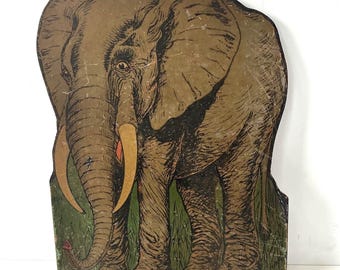 May include: Vintage wooden elephant cutout with a distressed finish. The elephant is depicted in shades of brown and tan, with long tusks and a red accent on its trunk. The base features green grass and a red border.