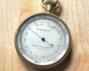 May include: A vintage brass barometer with a white face and black needle. The face has multiple scales and the text "OSKAR QUEISSER. MUNCHEN." The barometer is on a wooden surface.