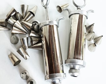 May include: Two silver cake decorating tools with a set of silver piping tips. The tools have a plunger and a nozzle for filling and decorating cakes.