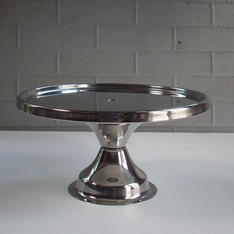 May include: A silver cake stand with a round, mirrored top and a flared base. The stand is made of metal and has a smooth, polished finish.