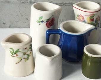 May include: A collection of six small ceramic creamers in various styles. One is solid olive green, one is solid white, one is deep blue with a handle, and three are white with floral patterns including red roses and white dogwood blossoms.