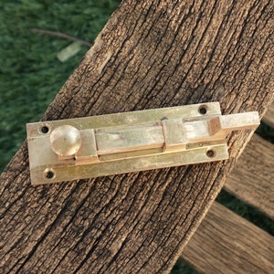 May include: A brass door bolt with a rounded knob and a sliding latch. The bolt is mounted on a weathered wooden surface.