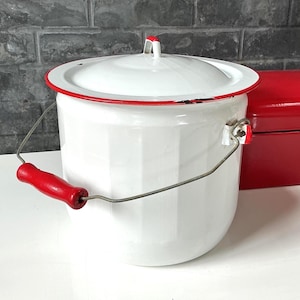 May include: A white enamel bucket with a matching lid, featuring a bright red rim and a wire handle with a red wooden grip. The cylindrical container has vertical ridges along its sides and a small red handle on top of the lid for easy removal.