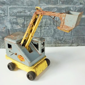 May include: A yellow and gray toy construction crane with a metal bucket. The crane has a yellow boom and a gray cab with the words "Toy Construction" printed on the side.