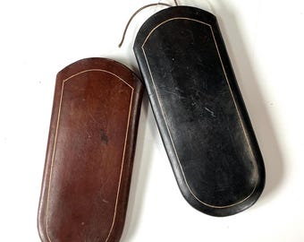 May include: Two eyeglass cases. One is dark brown, the other is black. Both have a rounded rectangular shape with a gold-colored trim. The cases are made of a leather-like material and are empty.