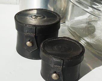 May include: Two small, cylindrical black leather containers with silver-coloured metal closures. The lids are embossed with a design. The containers are of different sizes and appear to be vintage or antique.