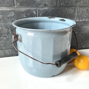 May include: A light blue enamel bucket with a vertical ribbed texture, a dark metal wire handle featuring a black wooden grip, and visible rust spots along the top rim. The bucket has a wide opening and a rounded base, showing signs of wear.