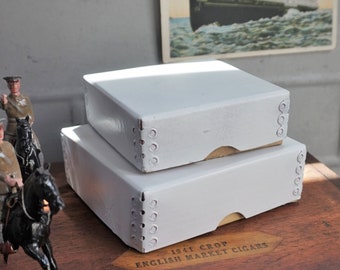 May include: Two white cardboard boxes stacked on top of each other. The top box is slightly smaller than the bottom box. The boxes have a decorative pattern of small circles around the edges. The boxes are sitting on a wooden surface with the text "1941 CROP ENGLISH MARKET CIGARS" printed on it.