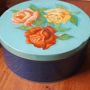 May include: A blue and white round tin with a floral design featuring three roses. The roses are red, orange, and yellow. The tin has a dark blue base.