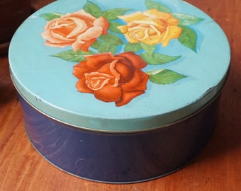 May include: A blue and white round tin with a floral design featuring three roses. The roses are red, orange, and yellow. The tin has a dark blue base.