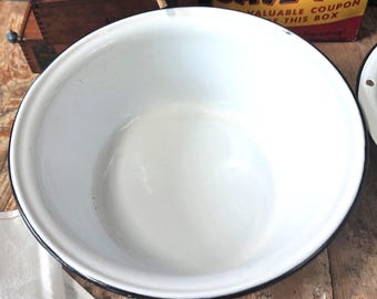May include: A white enamelware bowl with a black rim. The bowl is round and has a wide opening. A white pitcher is visible in the background, along with a vintage box with the text "SAVE 17¢".