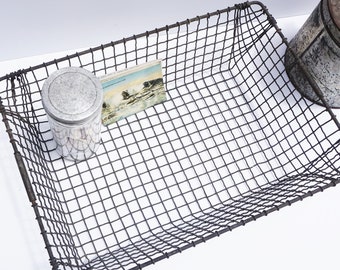 May include: A rectangular wire basket with a slightly curved bottom. The basket is made of dark grey metal wire and has a grid pattern. The basket is empty, but there is a small glass jar with a silver lid and a postcard inside.