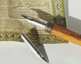 May include: A close-up of two vintage fountain pen nibs. The nibs are silver and have the words "JOSEPH GILLOTT" engraved on them. The nibs are on a white surface with a green and brown patterned paper in the background. The paper has the text "MANUFACTURED AT THE WORKS OF JOSEPH GILLOTT & SONS" printed on it.