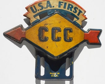 May include: Vintage metal sign featuring the text "U.S.A. FIRST" above a yellow diamond shape with "CCC". The sign has a blue and red arrow design and a blue bracket with "MADE IN U.S.A."