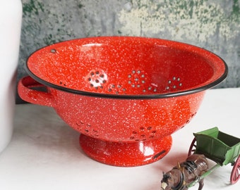 May include: A red enamel colander with a black rim and white speckles. The colander has a handle on the side and a base with holes.