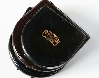 May include: A black, stacked, round, plastic case with a gold inscription that reads "Carl Zeiss Jena".